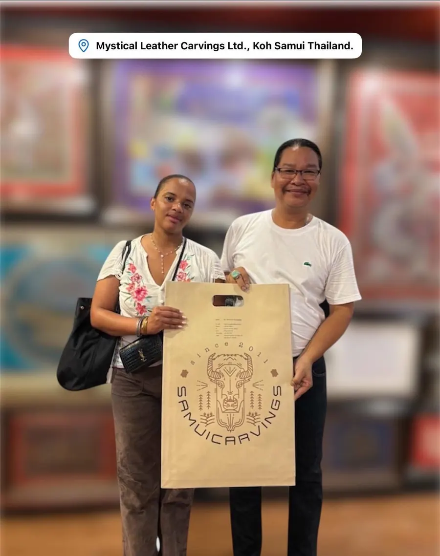 The artist with a guest holding a paper bag with the Samui Carvings logo, in his gallery